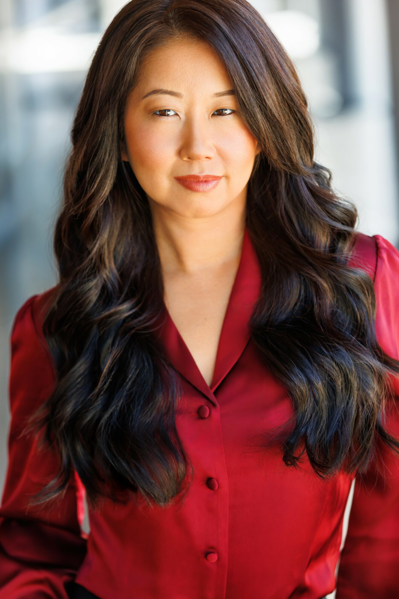 Woman with long dark hair wearing a red blouse, looking to the side, indoors with blurred background.