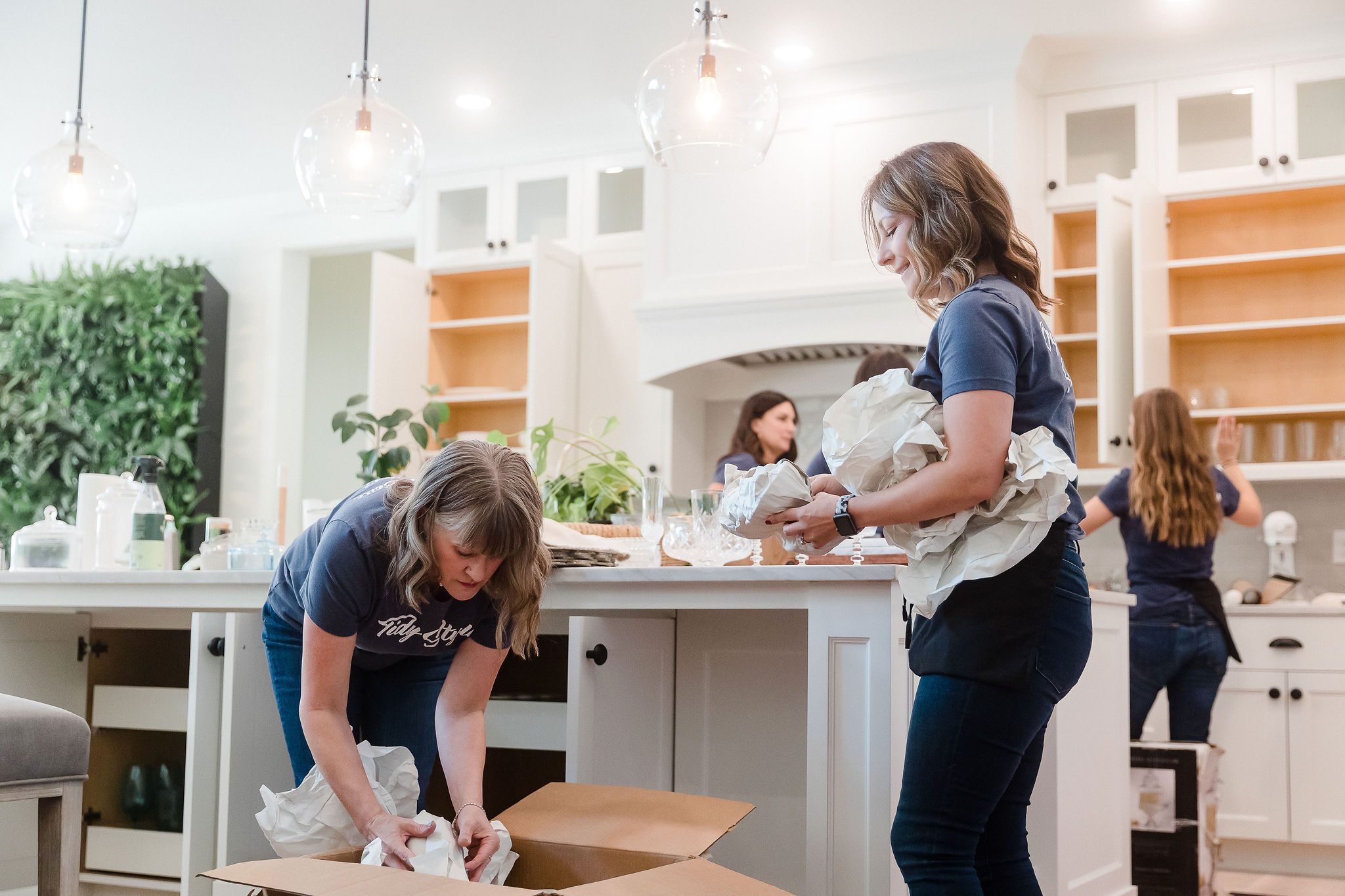 Four children in a bright kitchen, two cleaning, one reading, and one facing away, with white cabinets and greenery.