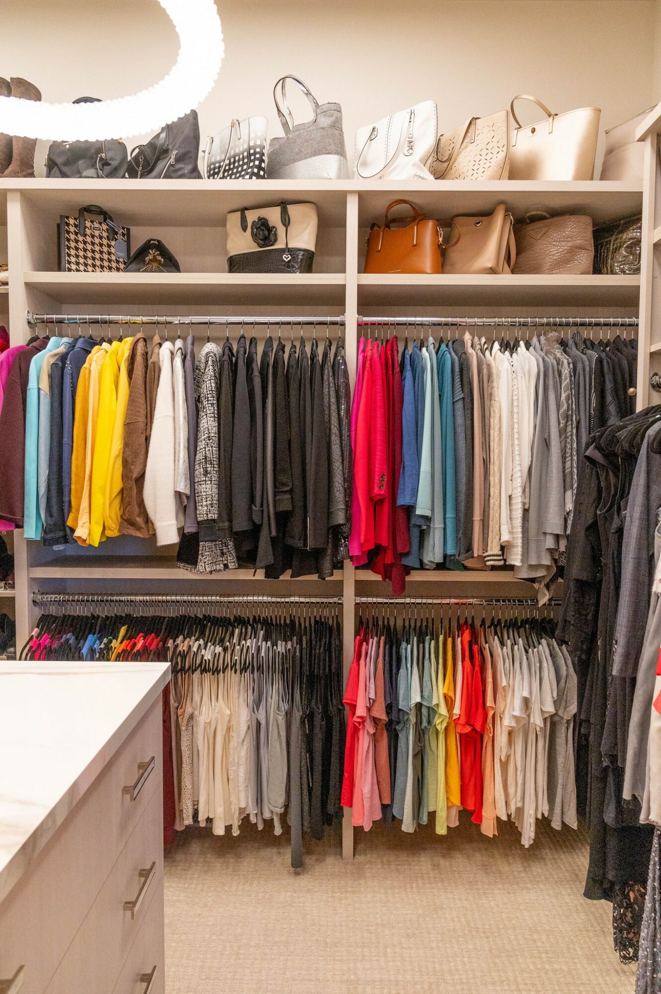 Closet with shelves of handbags and clothing racks with colorful shirts and pants, beige carpeted floor.