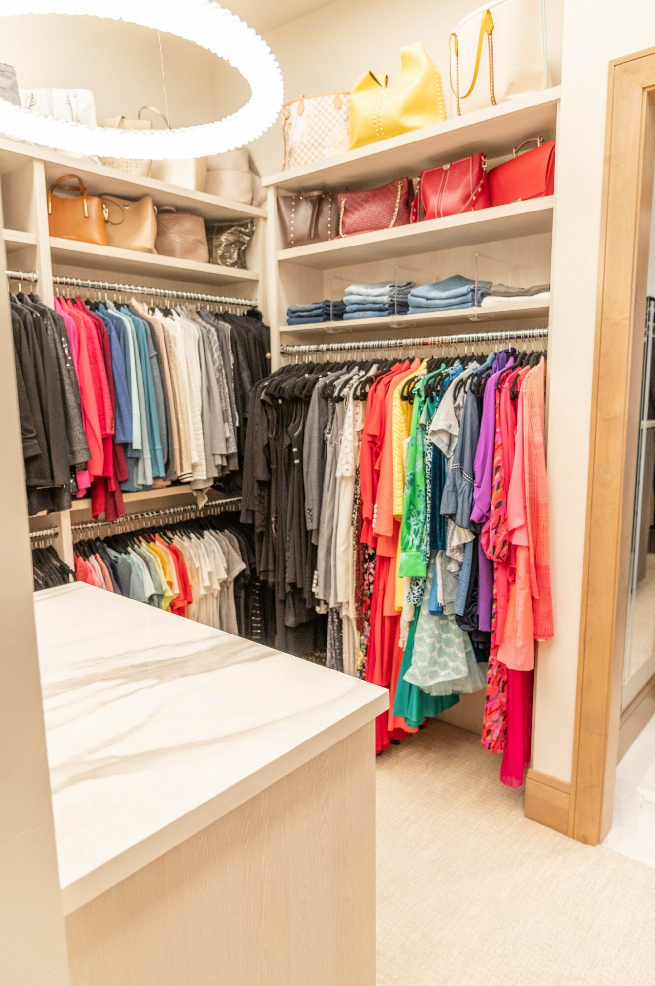 Closet with shelves of handbags, folded clothes, and hanging colorful garments, with a white marble-topped table in the foreground.