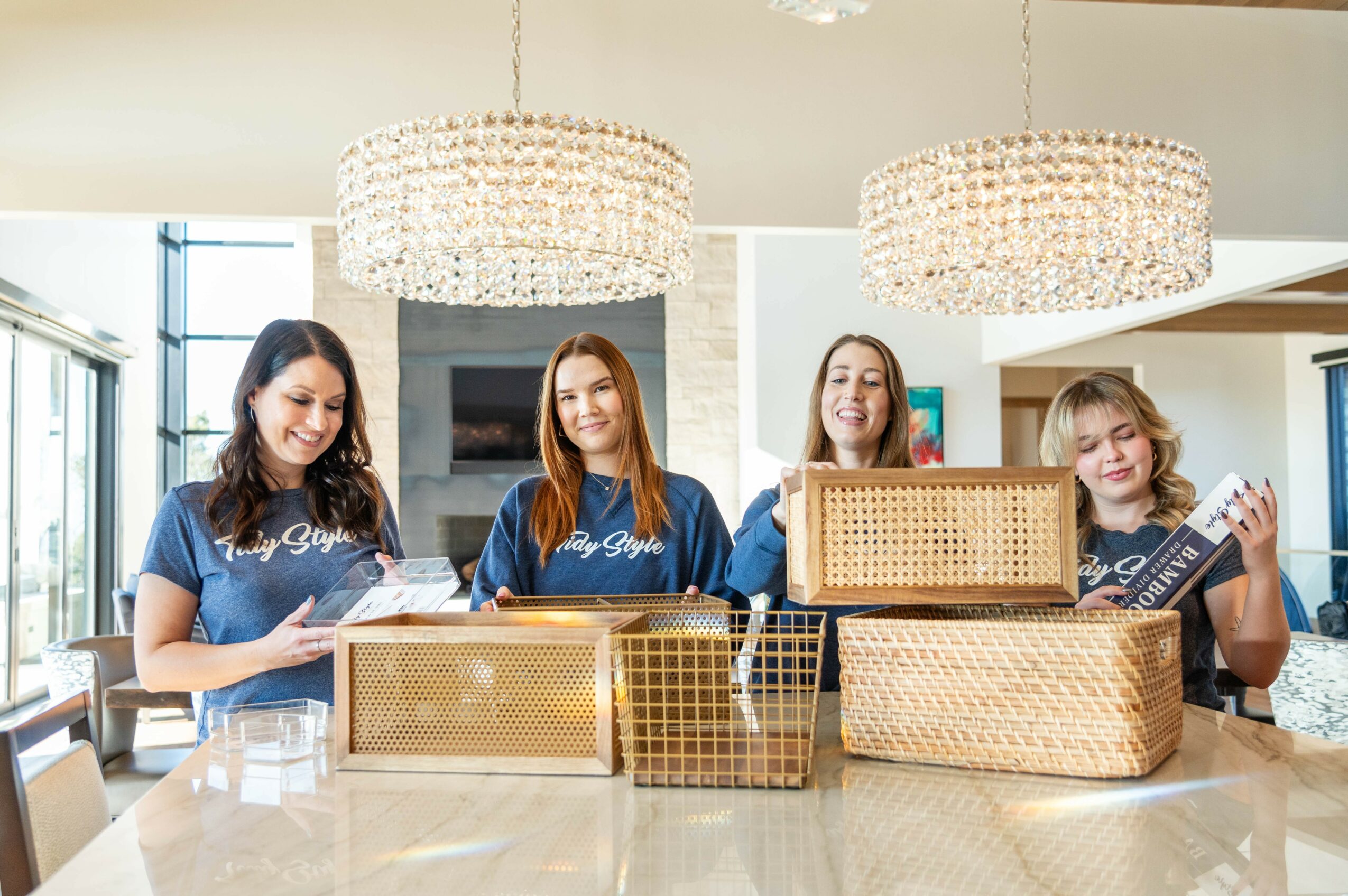Four women stand behind a table with woven baskets, smiling in a bright room with large chandeliers and windows.