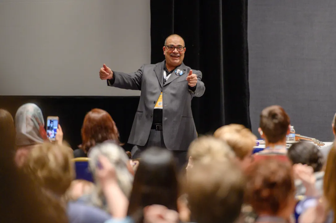 Man in gray suit speaking to an audience in a conference room, gesturing with hands, audience members taking photos.