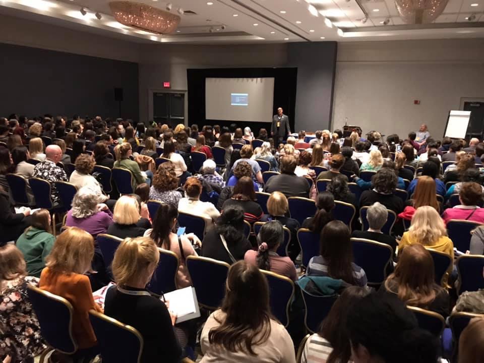 Large conference room filled with seated audience watching a presenter on stage with a screen, ceiling lights, and a whiteboard, 20 words.