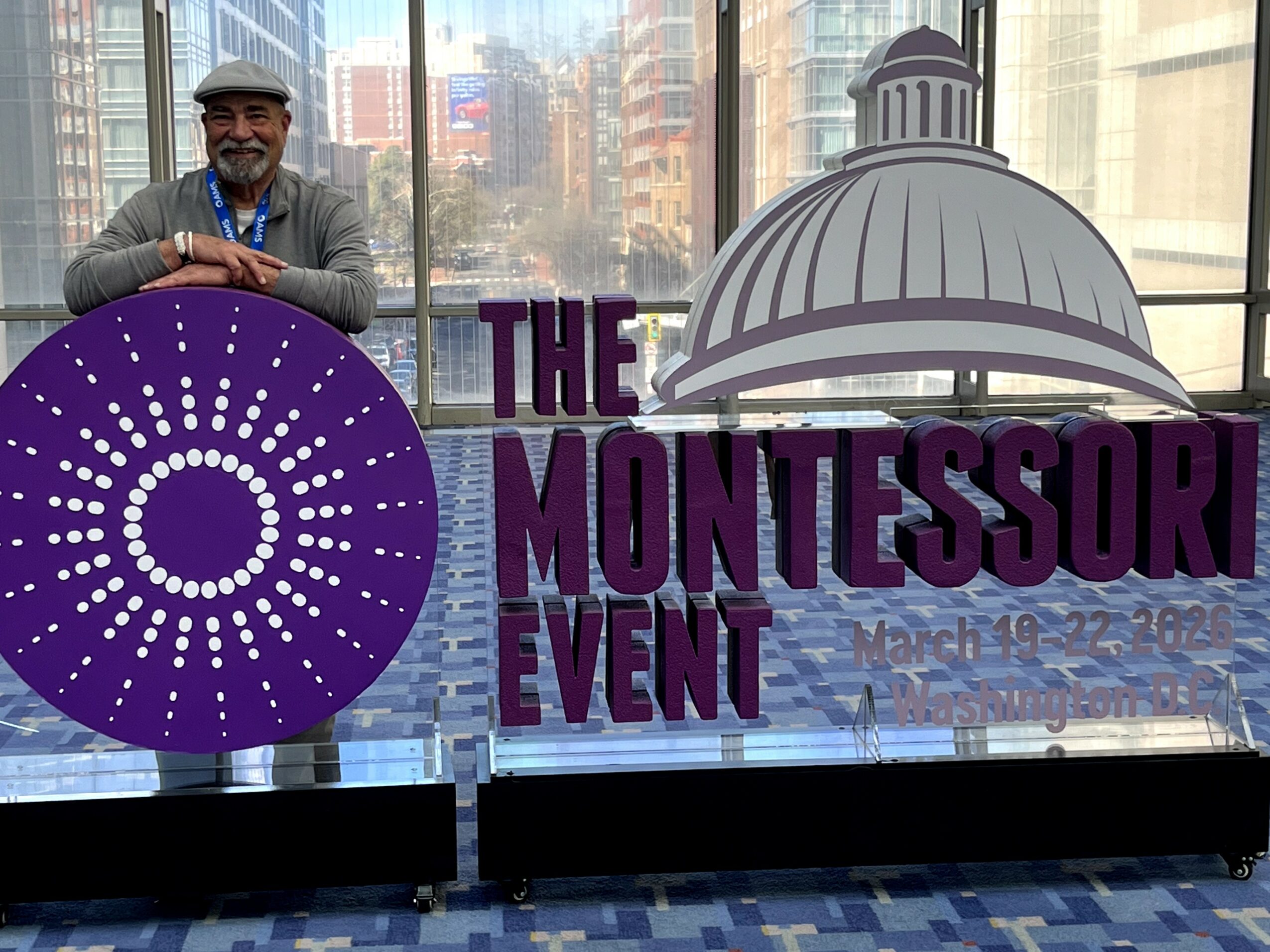 Person standing behind a purple circular logo with white dots, in front of a window with cityscape view, and a sign reading 'The Montessori Event' with a dome illustration.