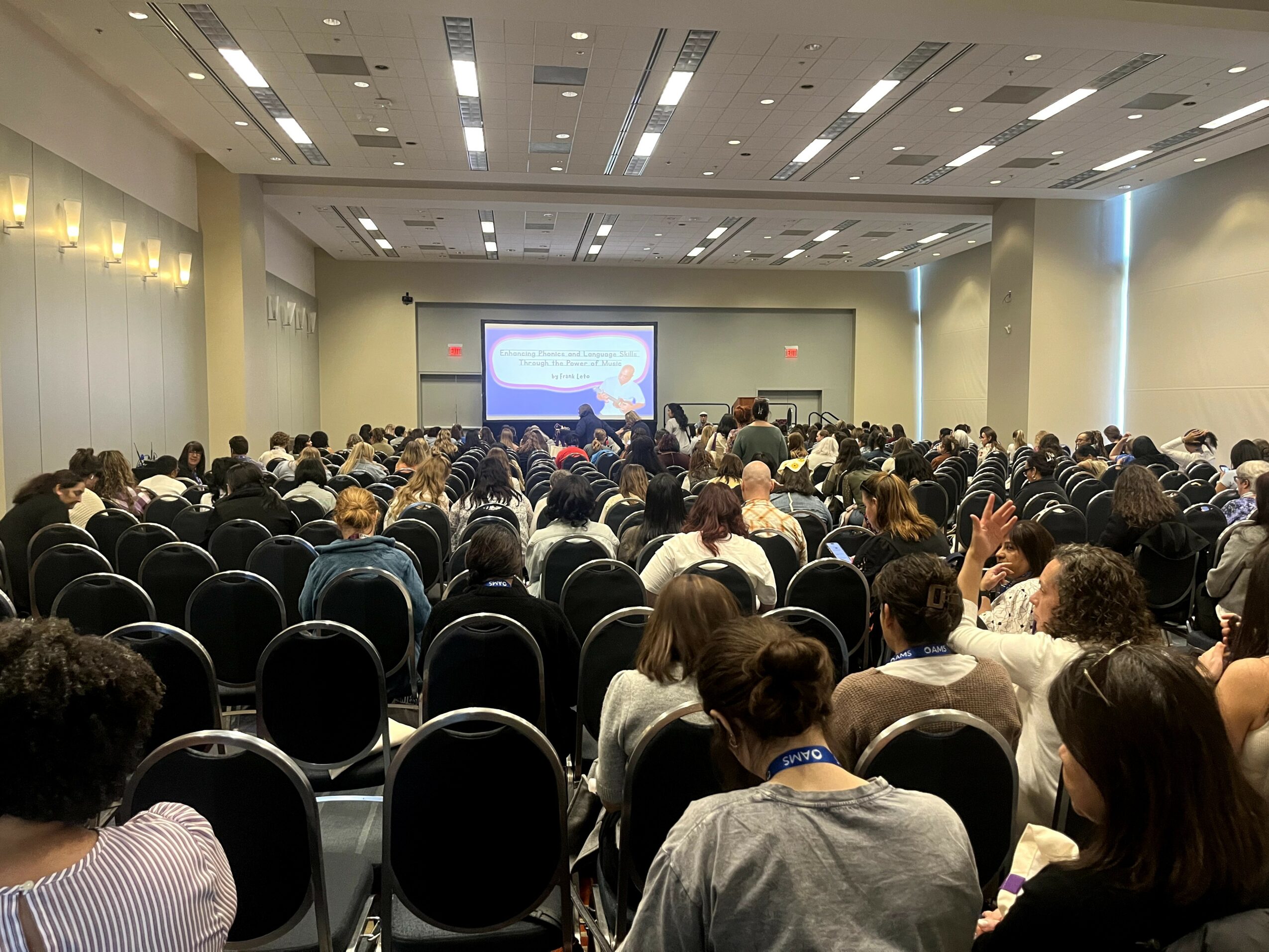Large conference room filled with seated audience facing a stage with a screen, ceiling lights, and walls, no visible text.
