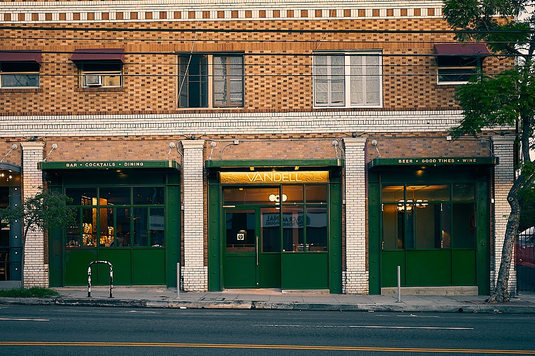Three storefronts with green doors and large windows on a brick building, street in front, trees on the side.