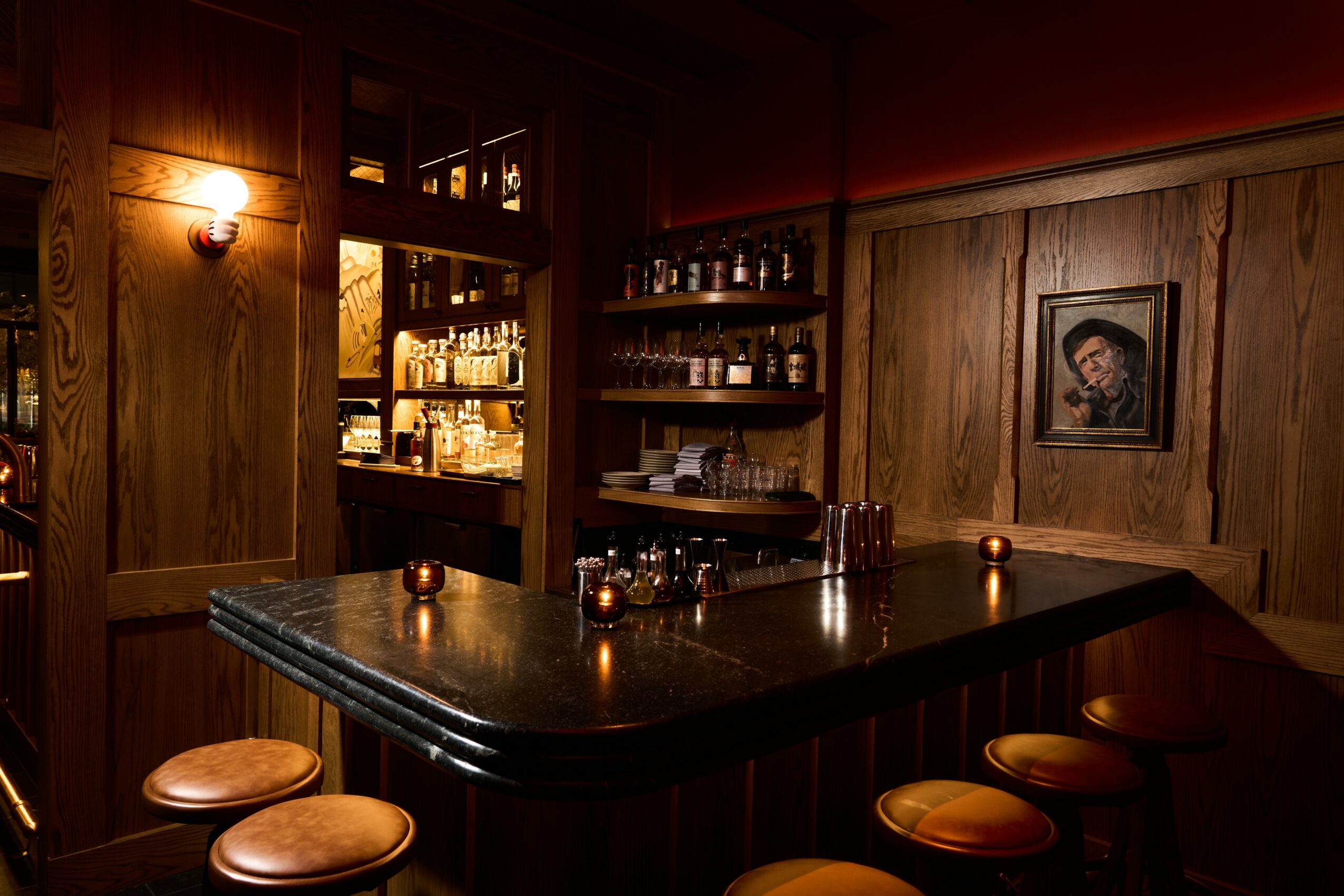 Bar with wooden paneling, shelves with bottles, and a framed picture on the wall, illuminated by warm lighting.