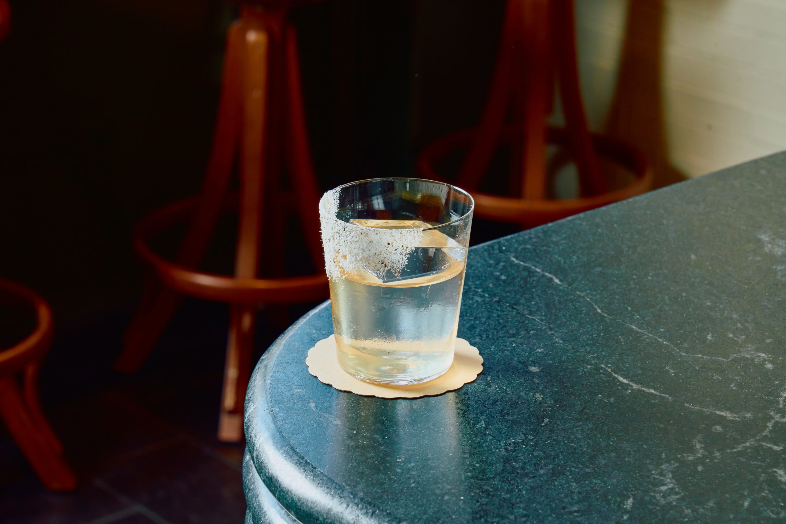 A glass of water on a coaster placed on a dark green marble table, with chairs in the background.
