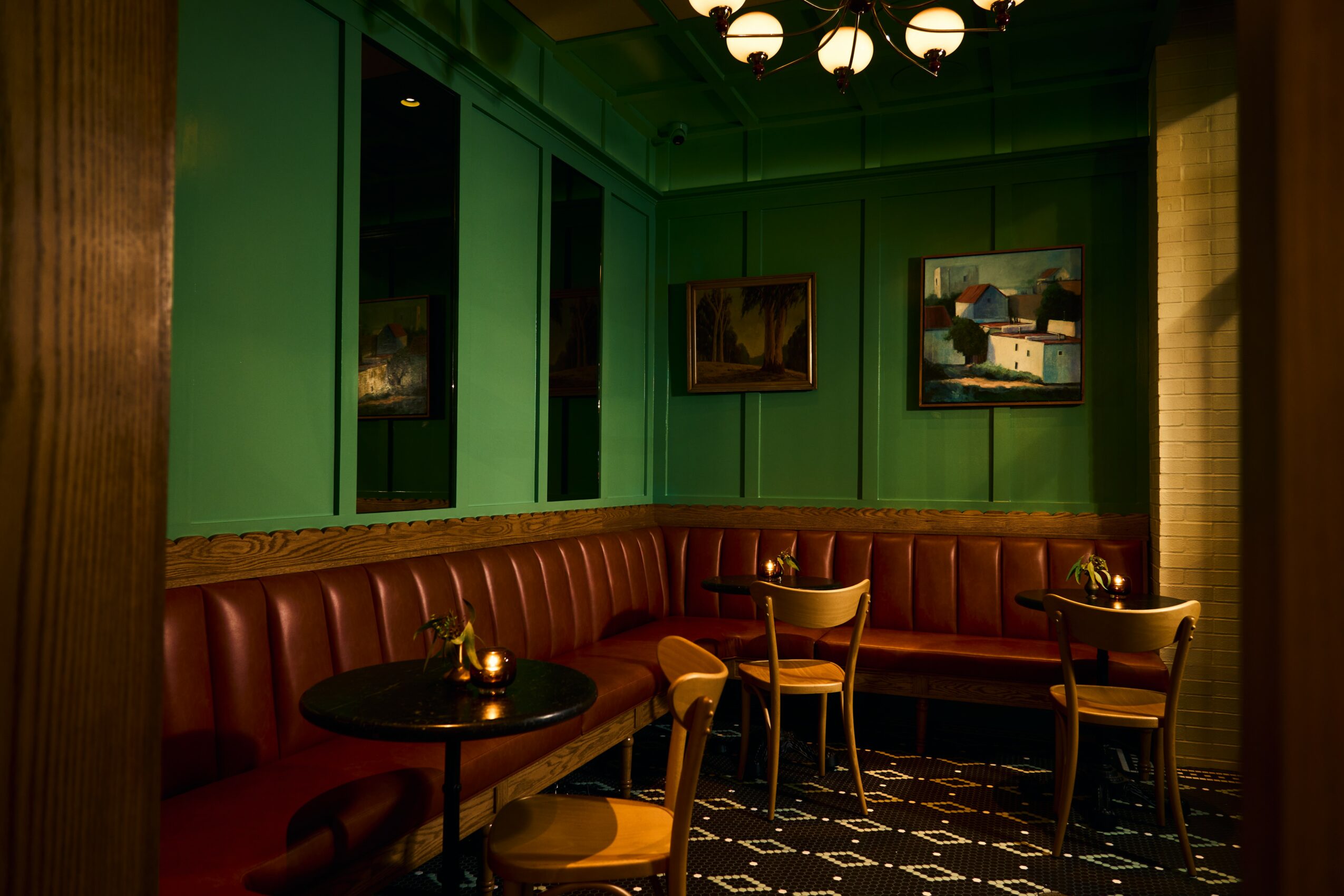 Interior of a restaurant with green walls, framed pictures, a curved leather banquette, and wooden chairs around tables.