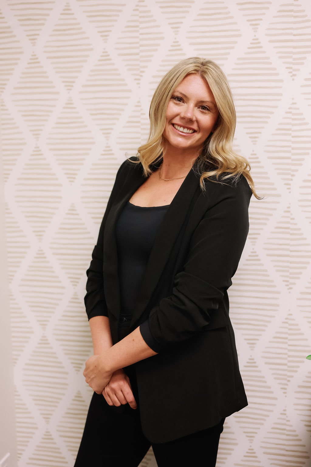 Woman with blonde hair smiling, wearing a black blazer and top, standing against a patterned background.