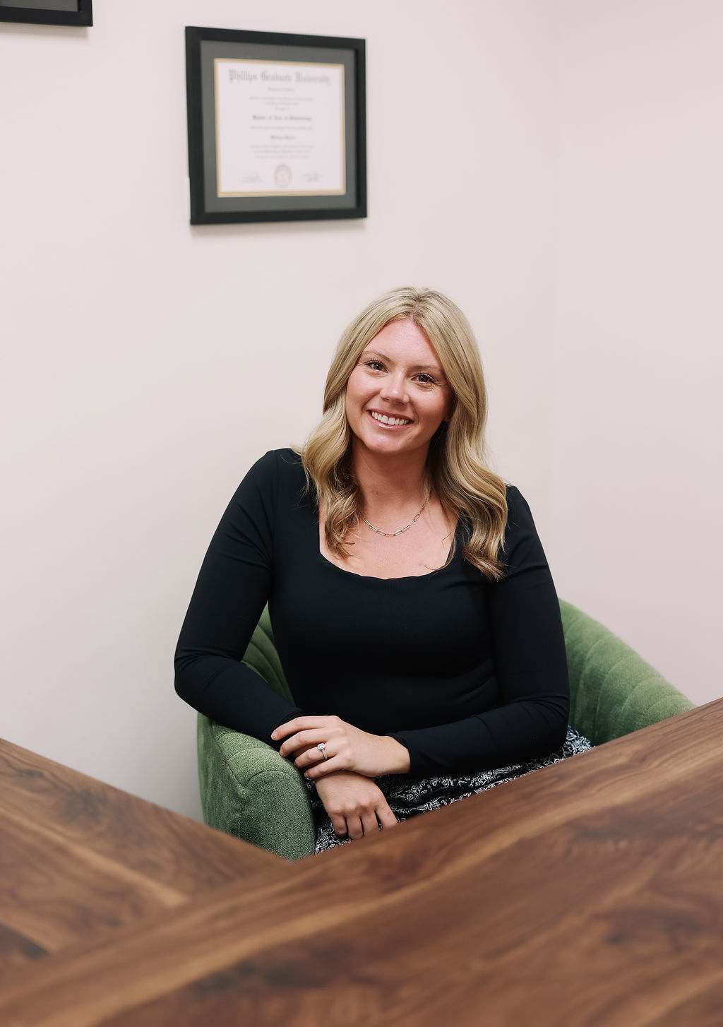 Woman with blonde hair smiling, sitting in a green chair, in an office with a framed certificate on the wall.