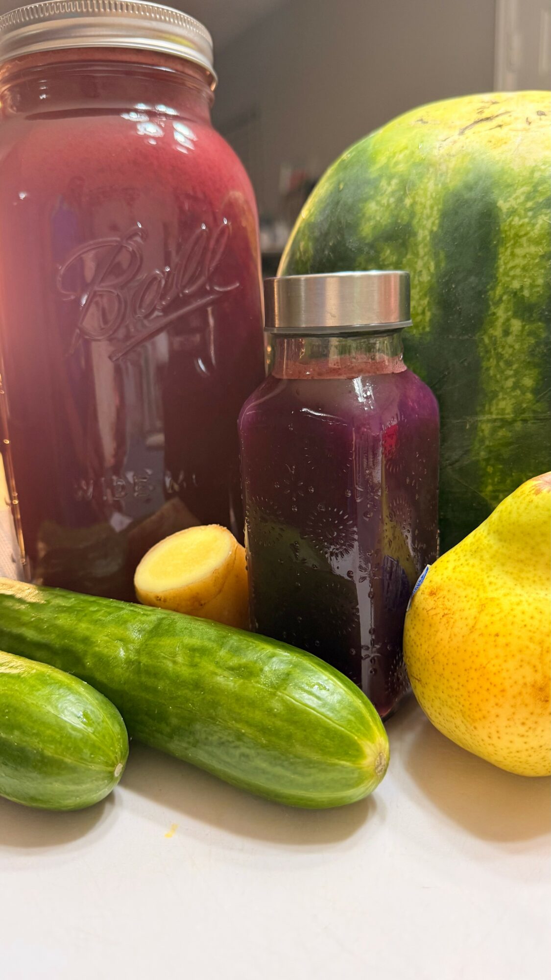 Watermelon, cucumber, lemon, and jars of purple juice on a white surface.