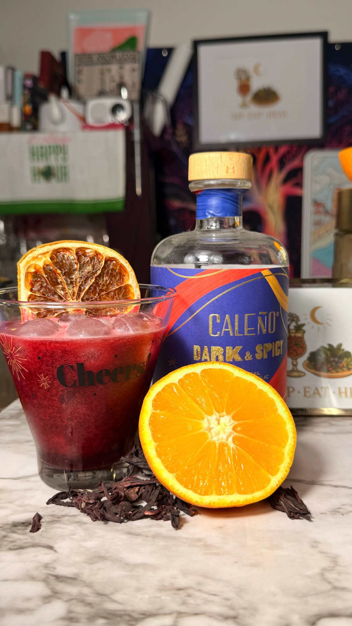 A glass with a dark red drink, orange slices, a bottle labeled 'Calenu Dark & Spice,' and an orange half on a marble surface.