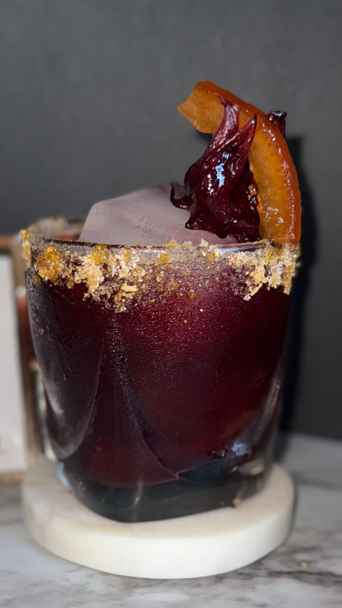 A cocktail in a glass garnished with orange and cherry slices, with a sugared rim, on a white coaster.