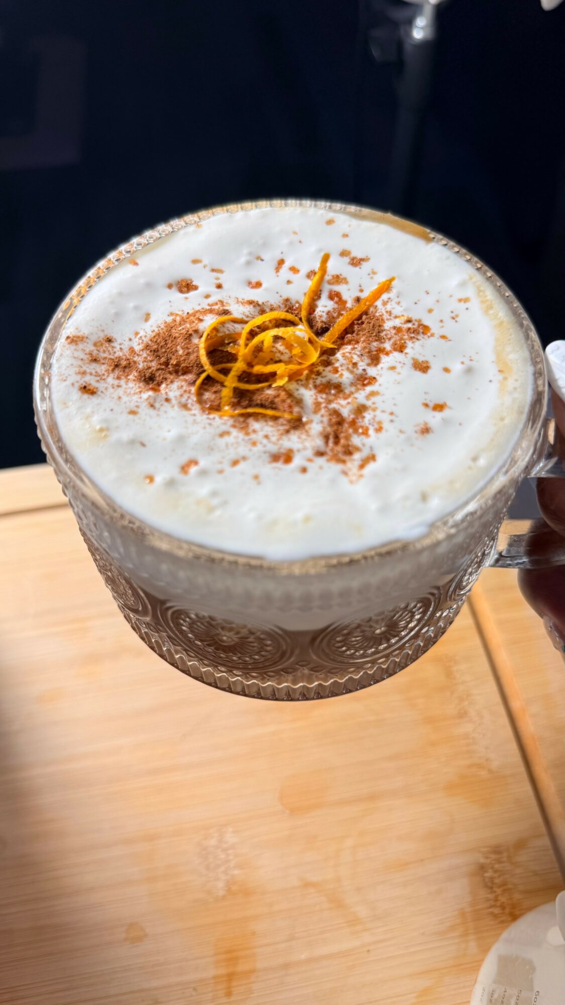 Foamy beverage in a glass with cinnamon and orange peel garnish, on a wooden surface.