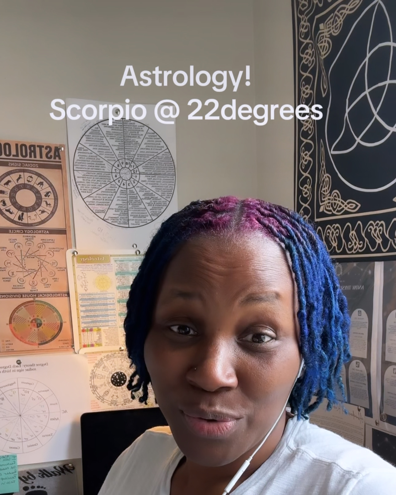 Person with colorful braided hair in a room with astrology posters and charts, wearing earphones, smiling slightly.