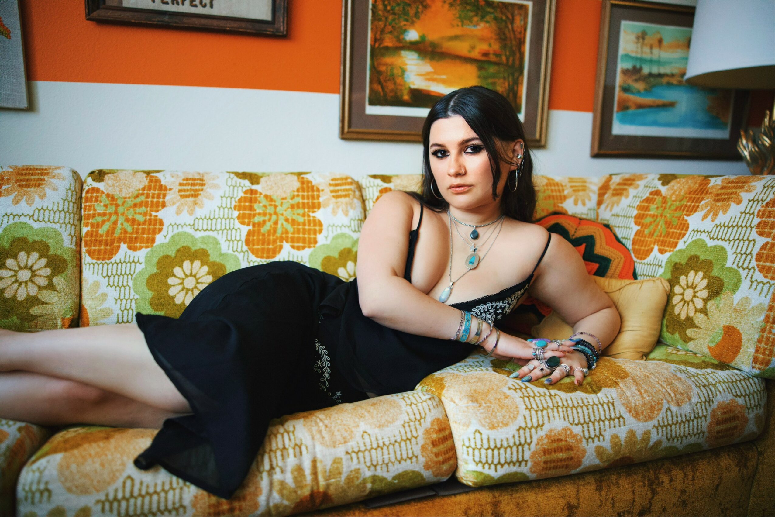 Woman with dark hair in a black dress lying on a colorful floral-patterned sofa, surrounded by framed artwork.