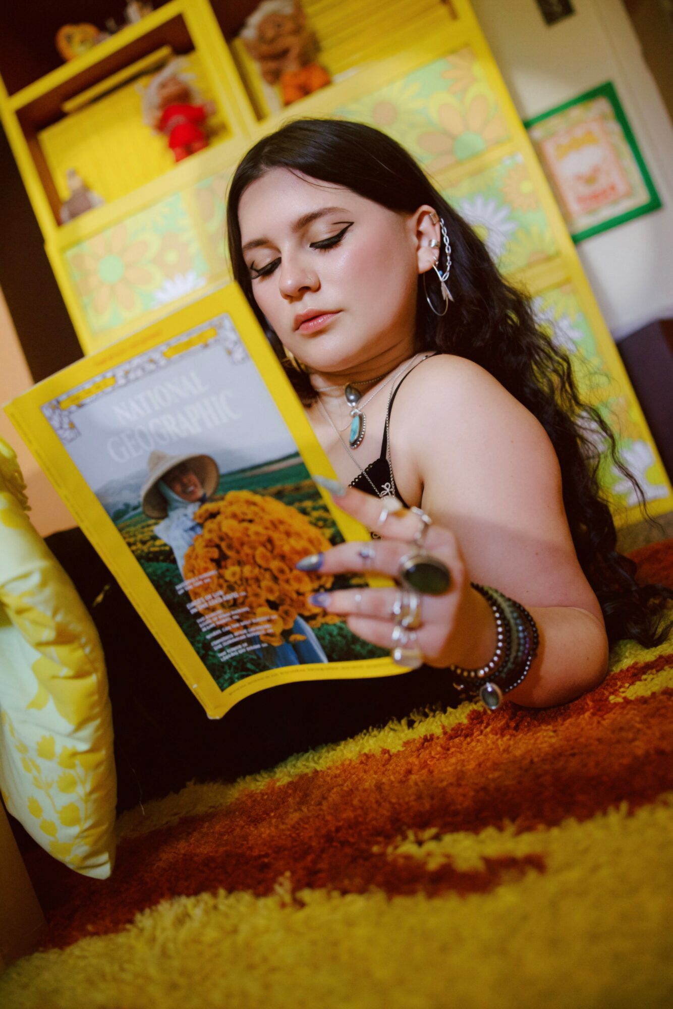 Young woman with dark hair and hoop earrings reads a magazine, sitting on a colorful carpet with yellow shelves in background.