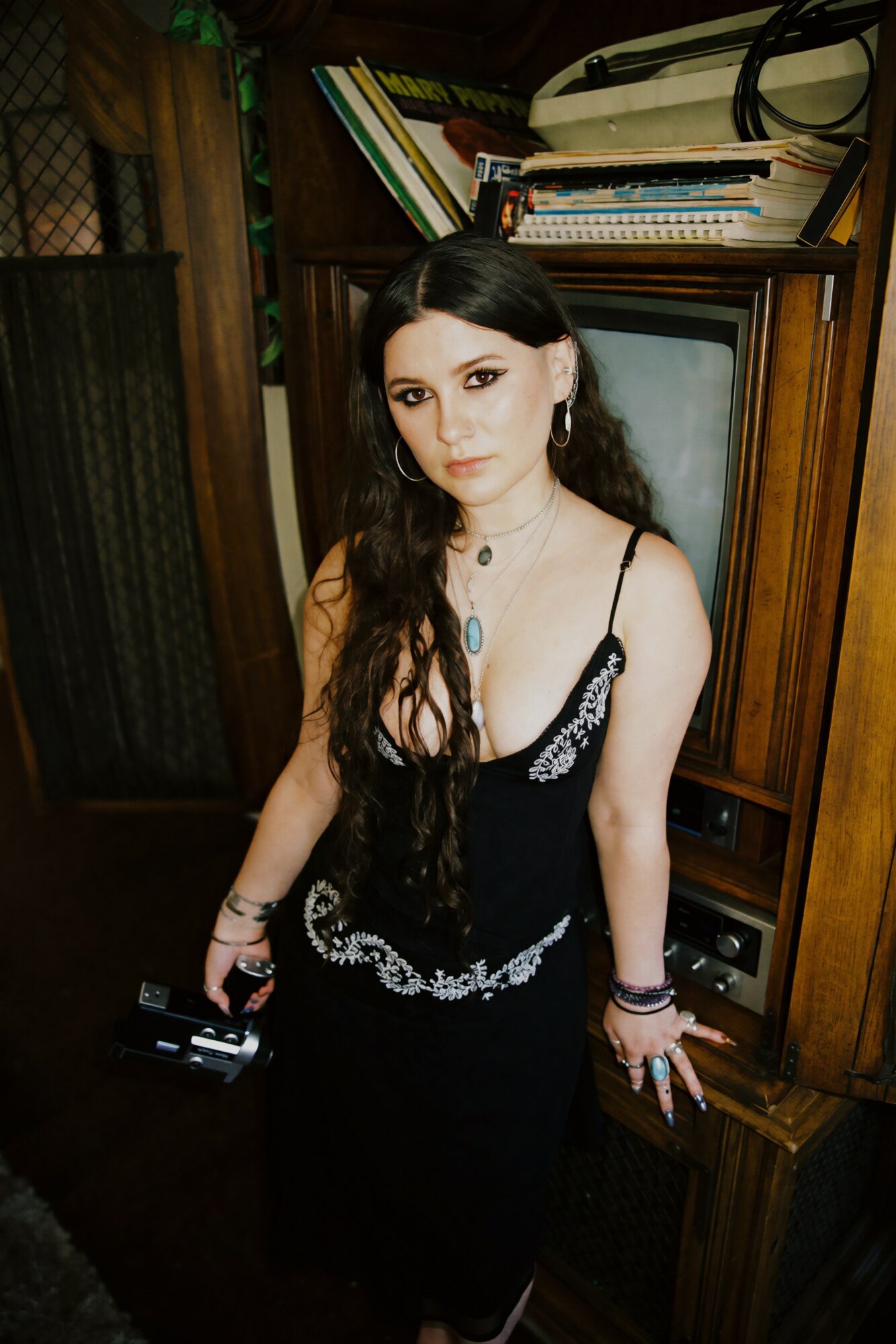 Young woman with long dark hair wearing a black dress with lace details, standing indoors near wooden furniture.
