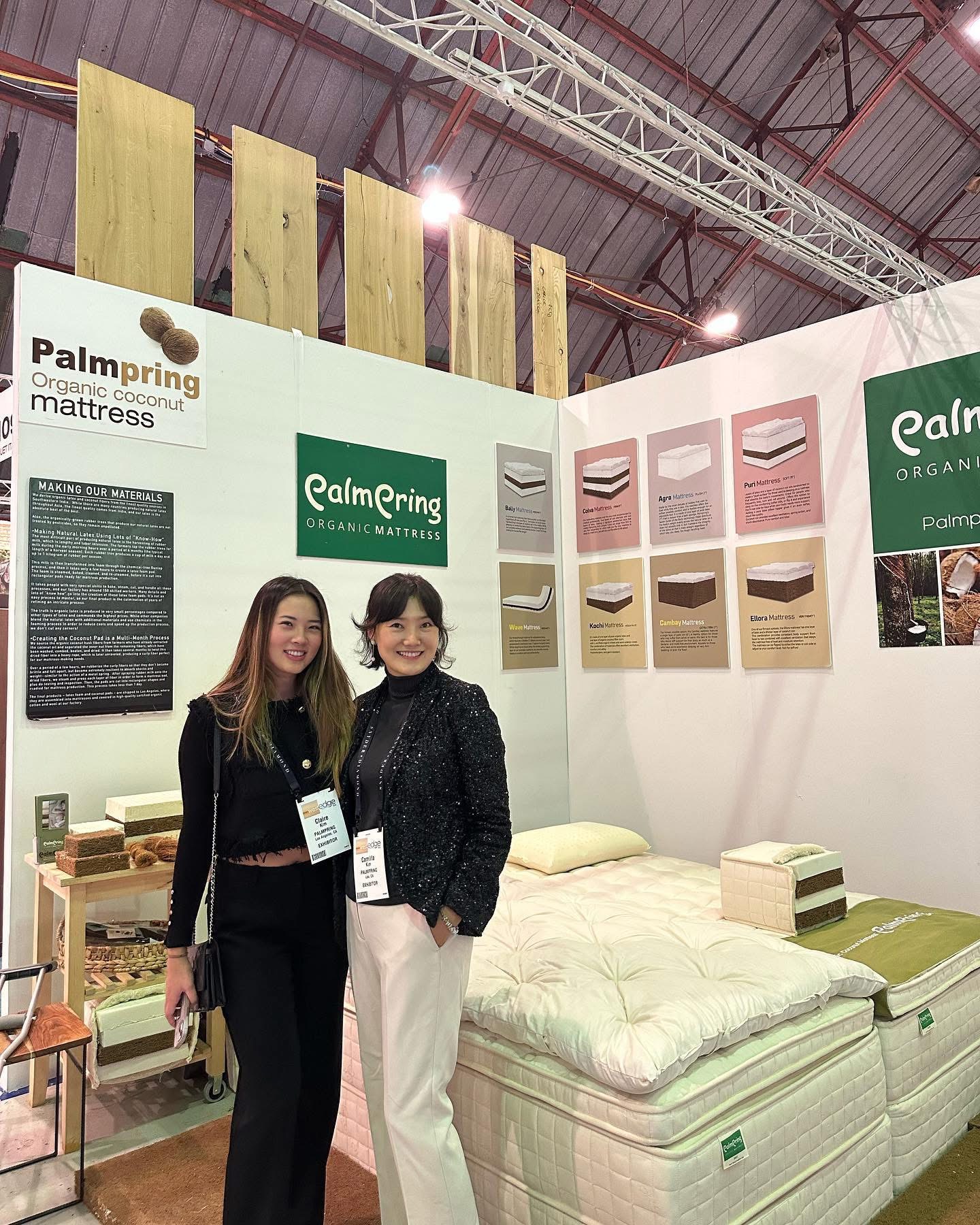 Two women standing in front of a display booth with mattresses and color samples at an exhibition, smiling at the camera.