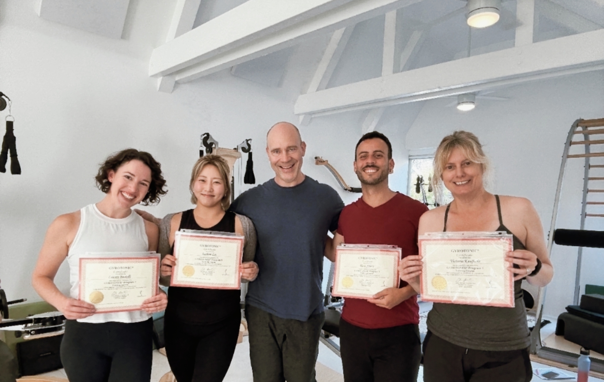 Group of five people holding certificates indoors, smiling, in a bright room with white walls and ceiling.