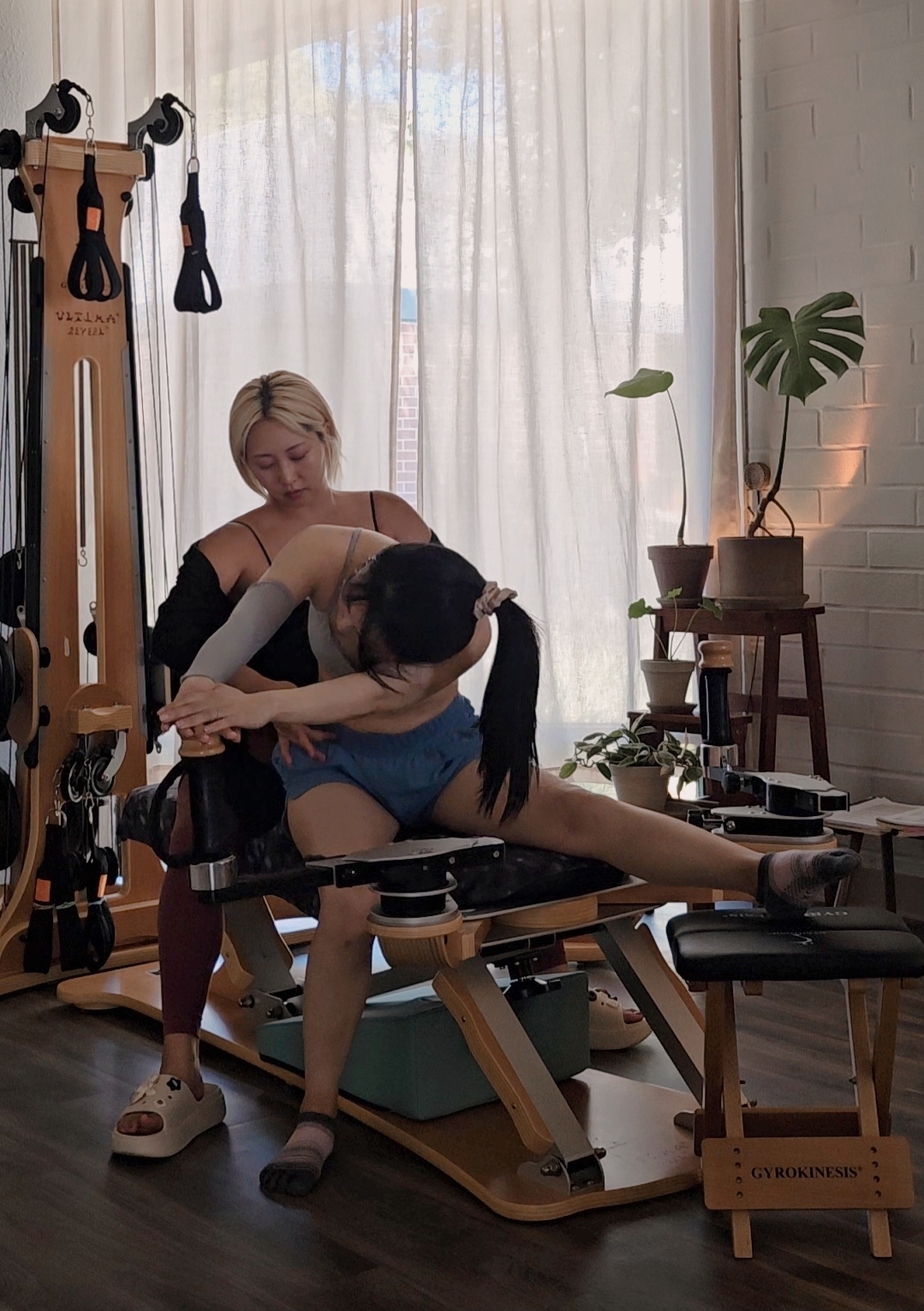 Two women in a room with large window and plants, one assisting the other with stretching on a massage table.
