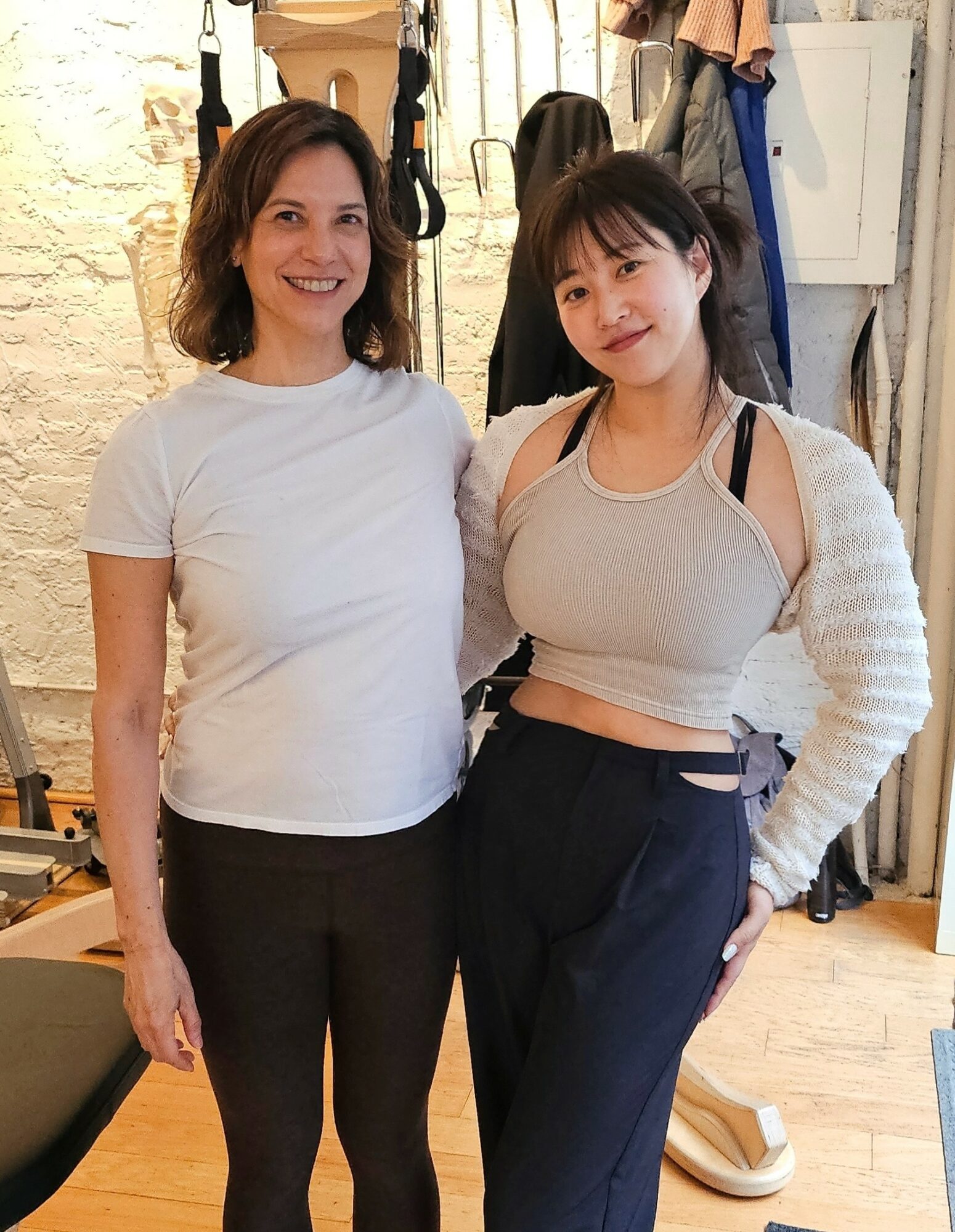 Two women standing indoors, smiling, with clothing and accessories in the background.