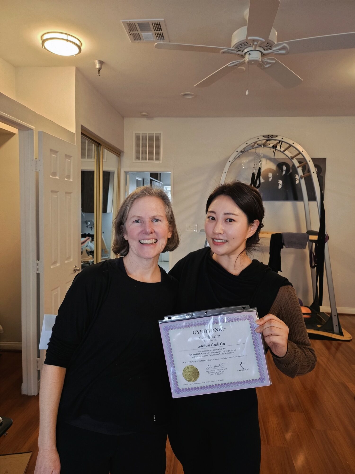 Two women standing indoors, one holding a certificate, smiling at the camera, with a ceiling fan and exercise equipment in the background.