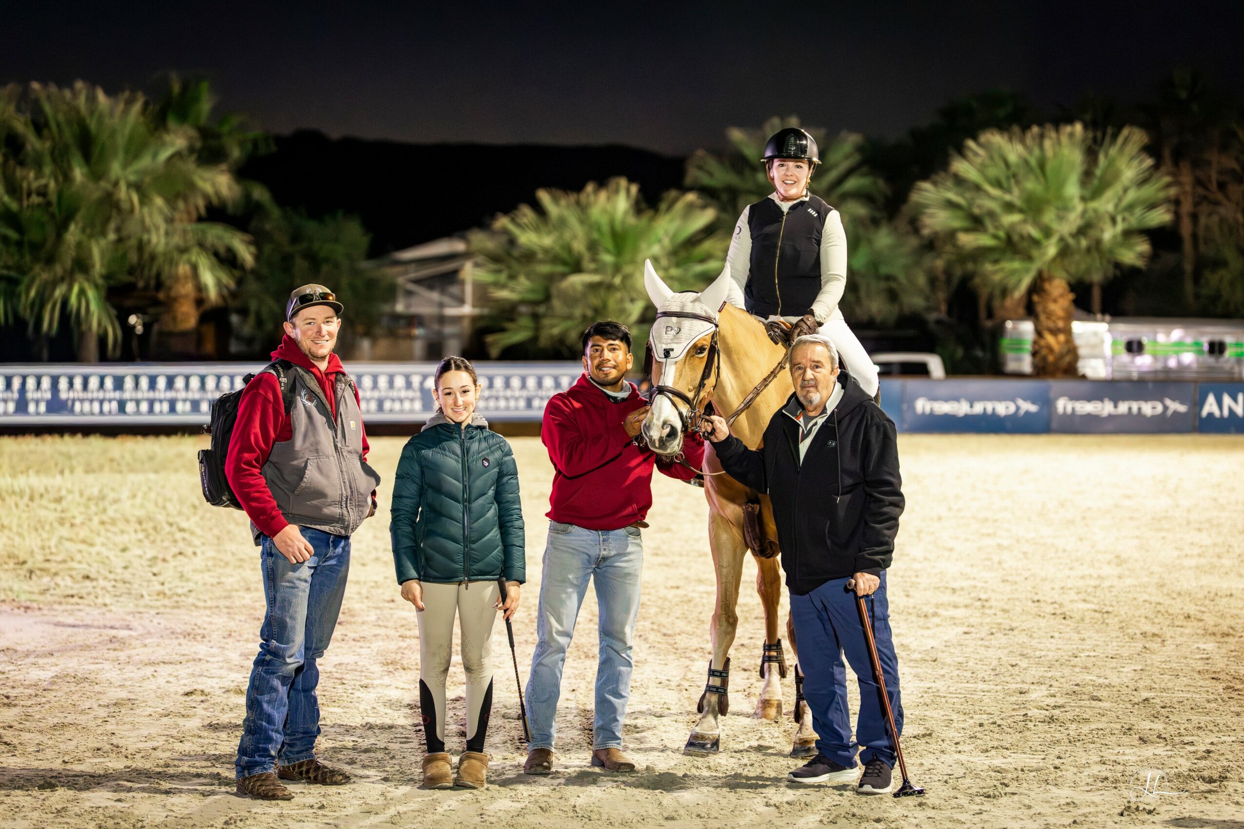 Four people and a horse in an outdoor arena at night, with palm trees in the background.