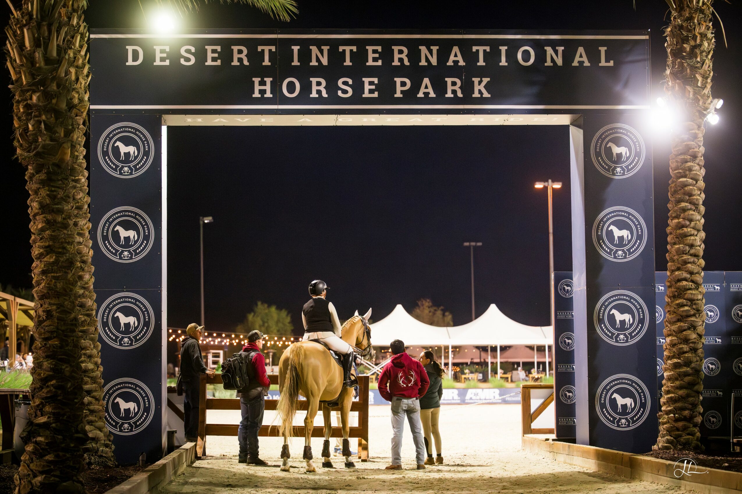 Night scene at horse park with people and a horse under a large archway, tents in background, lights illuminating the area.