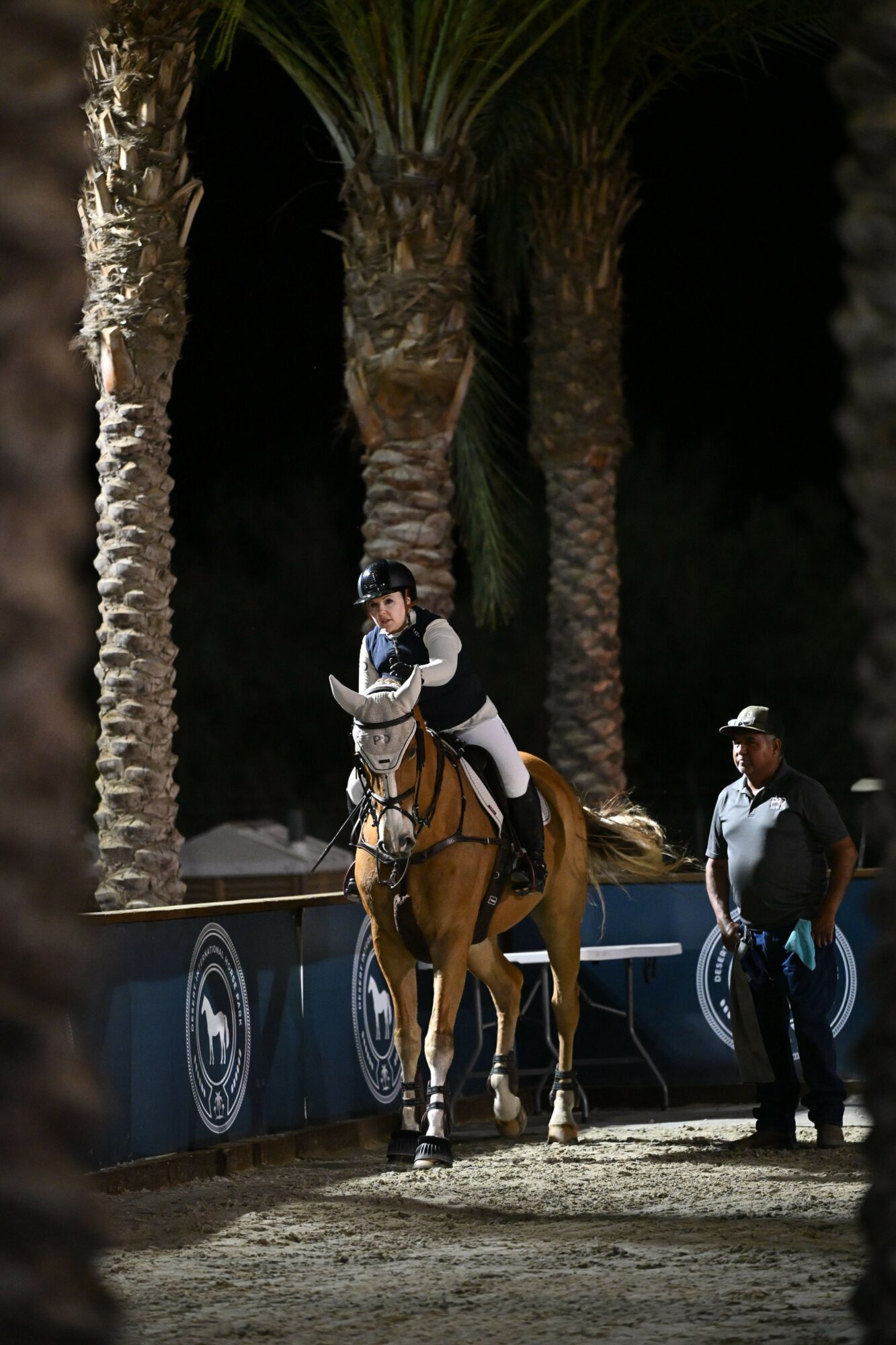 Two people standing outdoors near palm trees and a dark cave entrance, with a blue sign and a person in a brown coat.