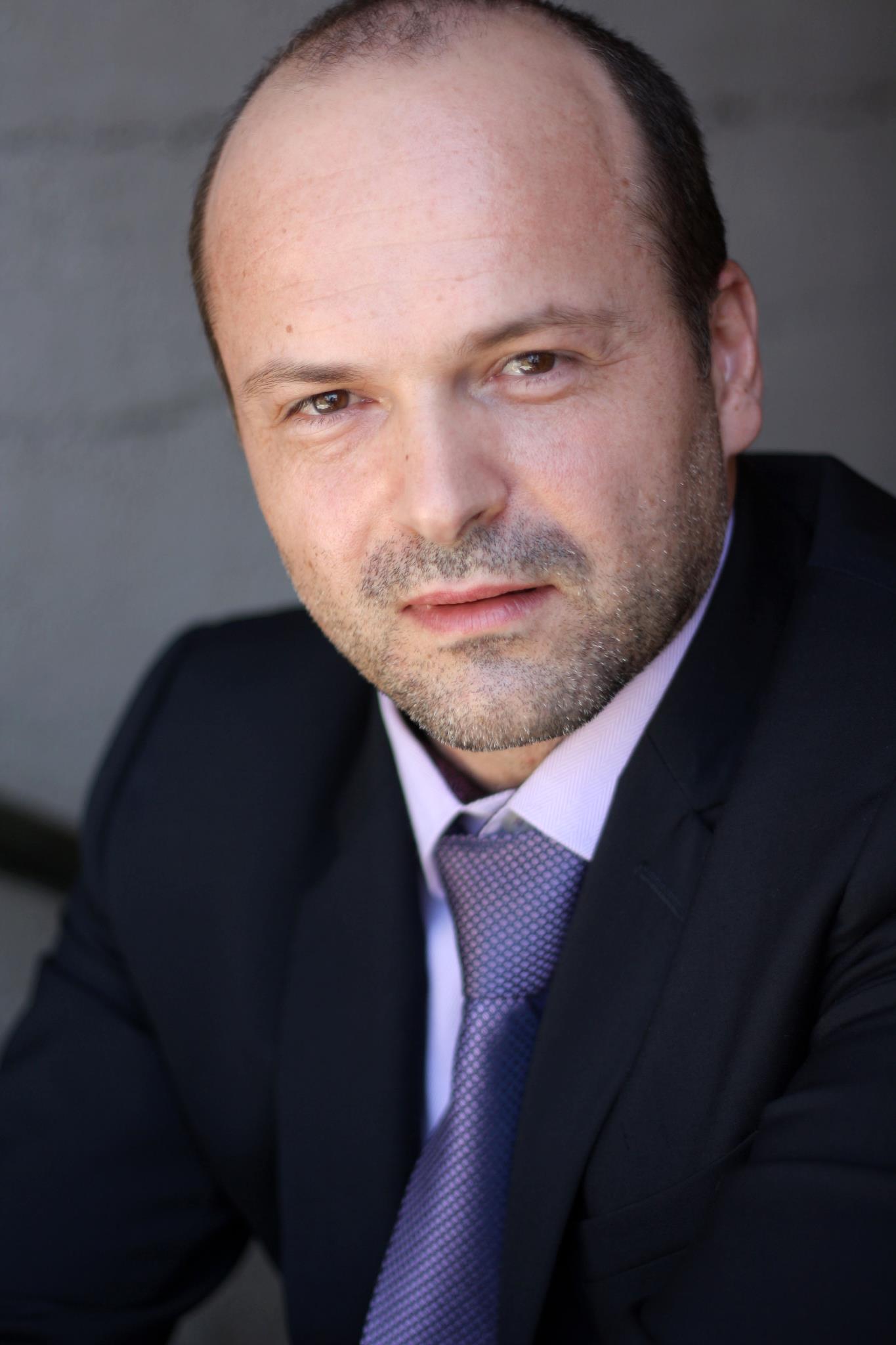 Man with a receding hairline and beard wearing a dark suit, white shirt, and purple tie, looking at the camera.