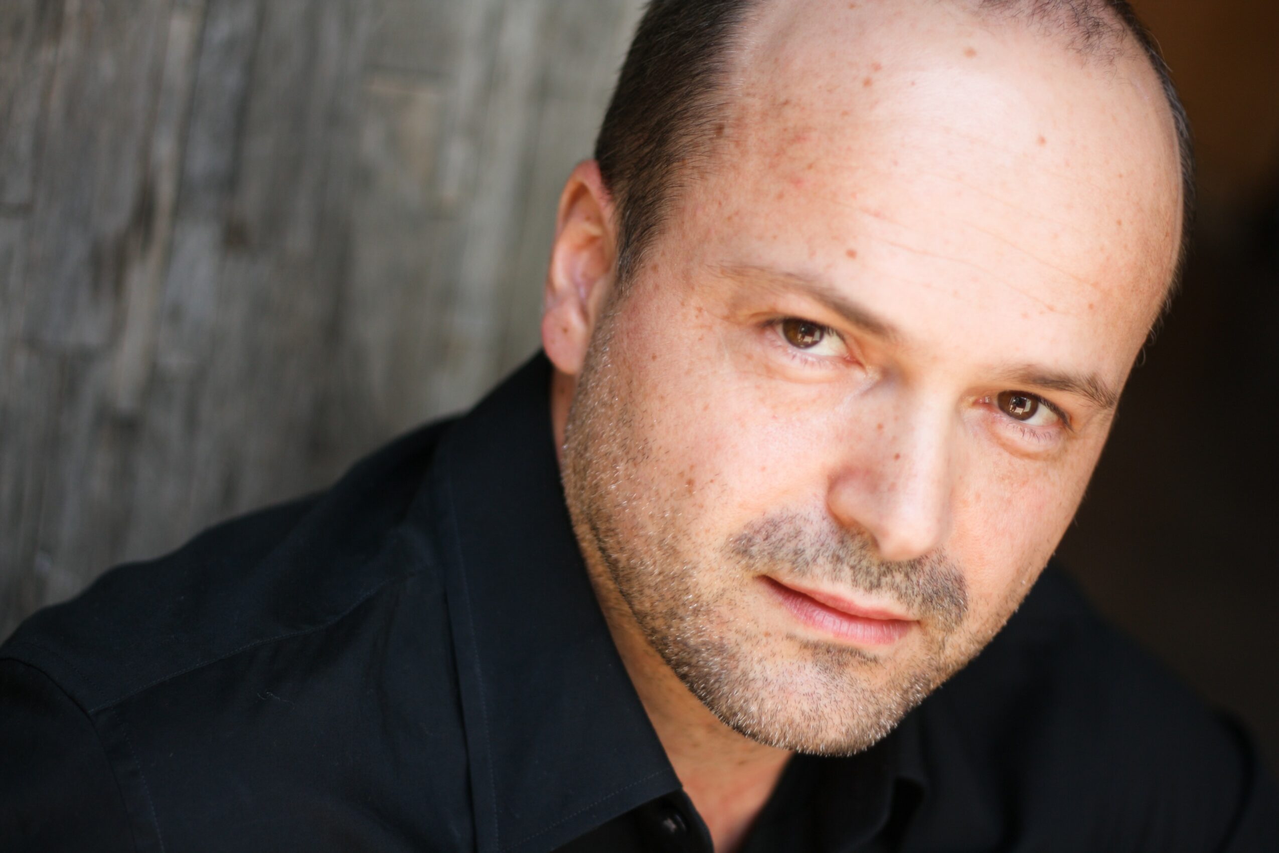 Close-up of a man with short hair and light stubble, wearing a black shirt, looking at the camera.