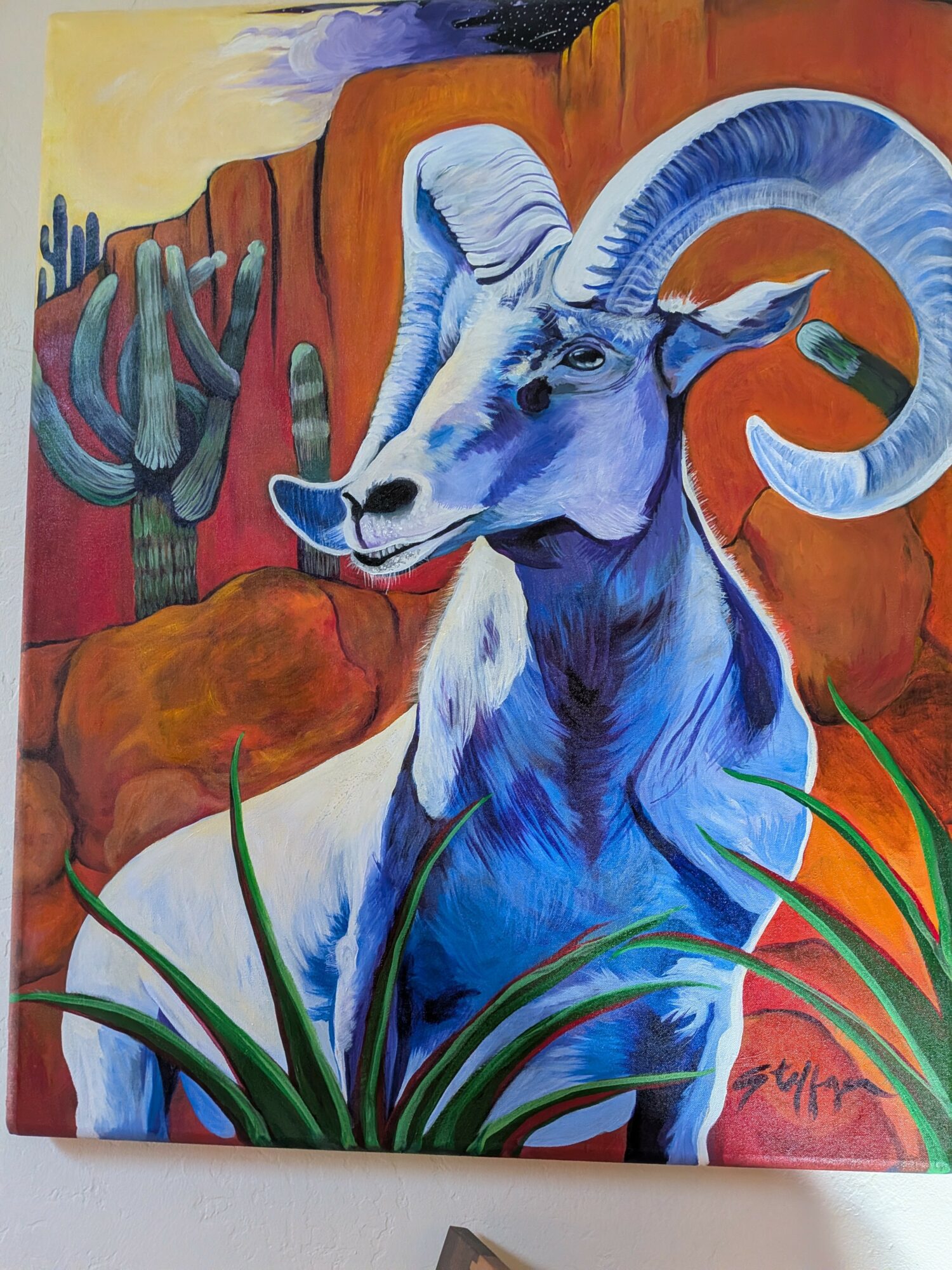 Blue and white goat with large curved horns in a desert landscape with cacti and red rock formations.