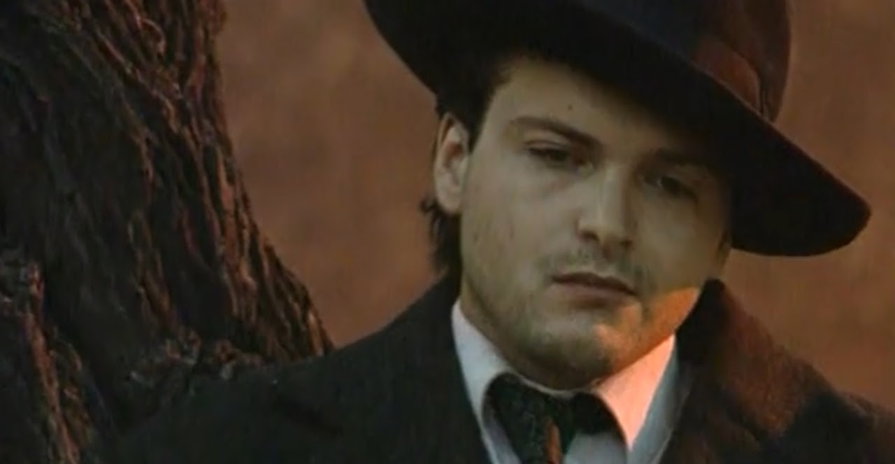Young man wearing a hat and dark coat, standing outdoors near a tree, with a serious expression.