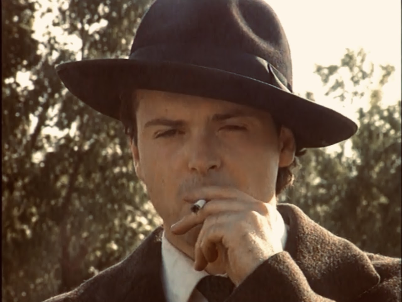 Man wearing a wide-brimmed hat, smoking a cigarette, outdoors with trees in background.