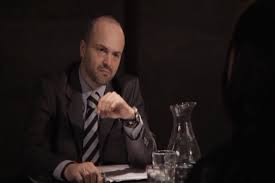 Man with a beard and suit sitting at a table with a glass pitcher and glass, looking at the camera.
