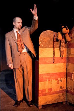Man in a suit standing on stage, waving, with a wooden puppet on a stand beside him, against a black background.