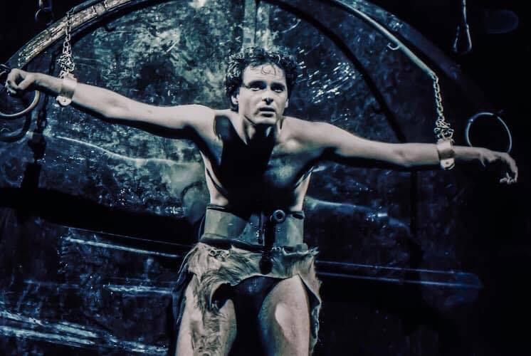 Person with arms outstretched, sitting on a chair, in a dark, theatrical setting with chains and a reflective background.