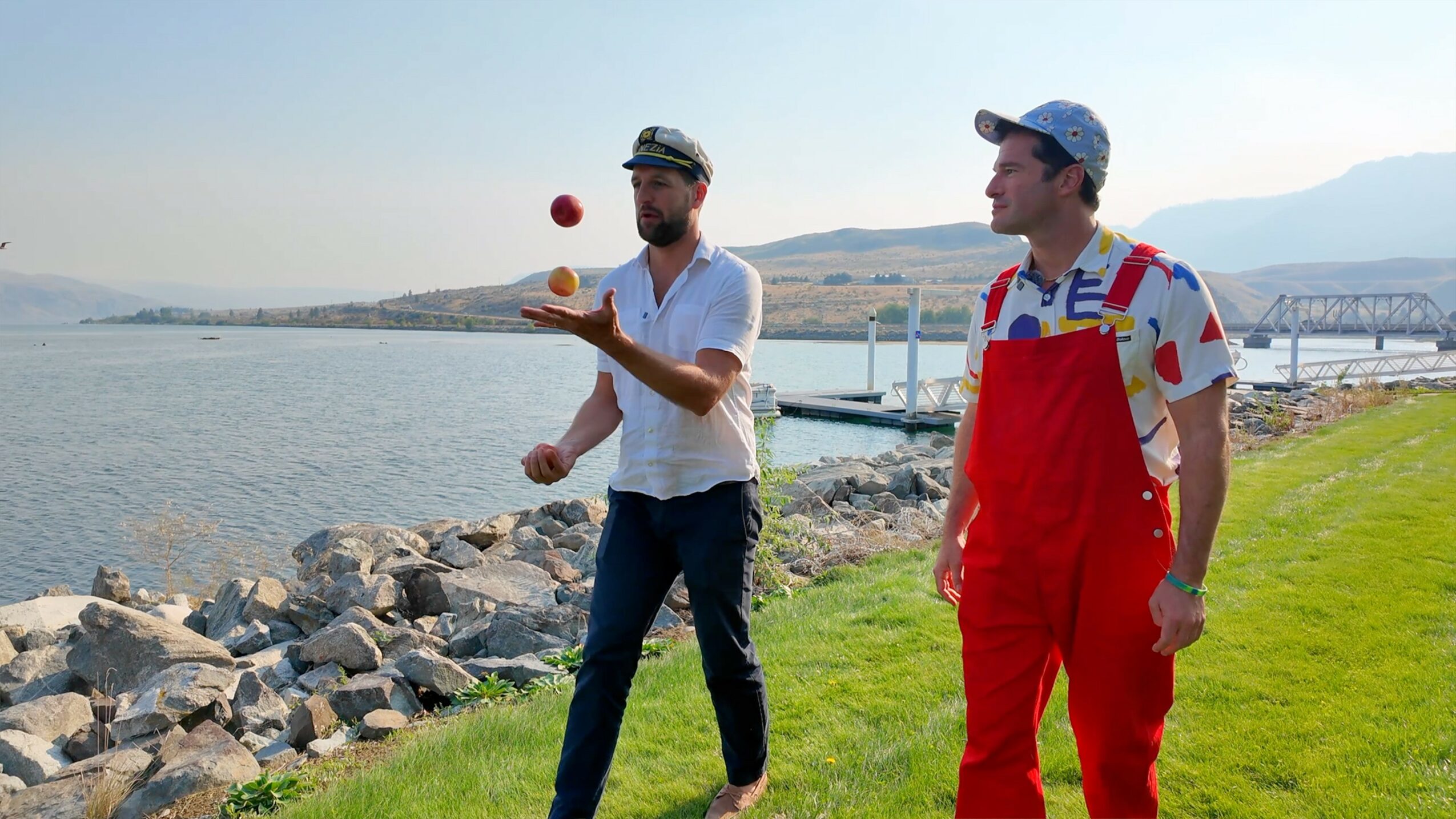 Two men stand outdoors near water, one juggling a red ball, the other watching, with a rocky shoreline and bridge in background.