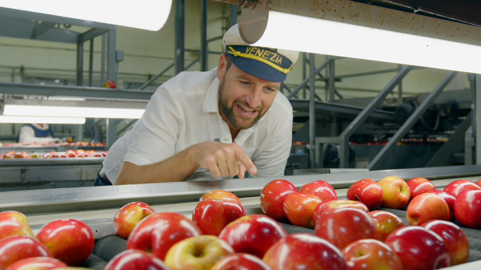 Man inspecting apples in a factory, smiling, with apples in foreground and industrial equipment in background.