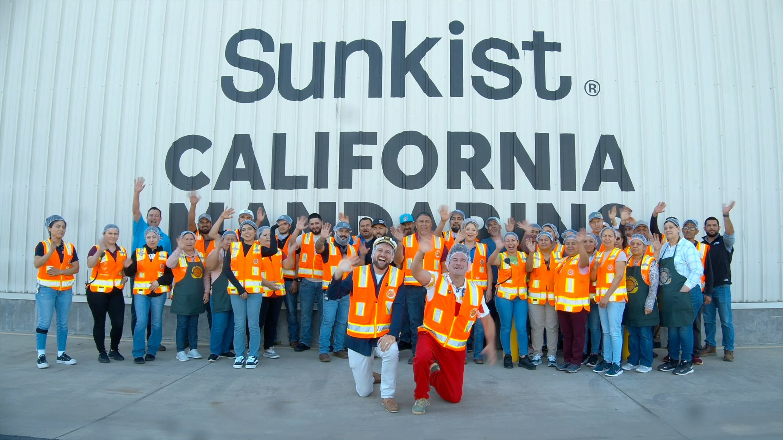 Group of people wearing orange safety vests standing in front of a large wall with 'Sunkist CALIFORNIA' text.