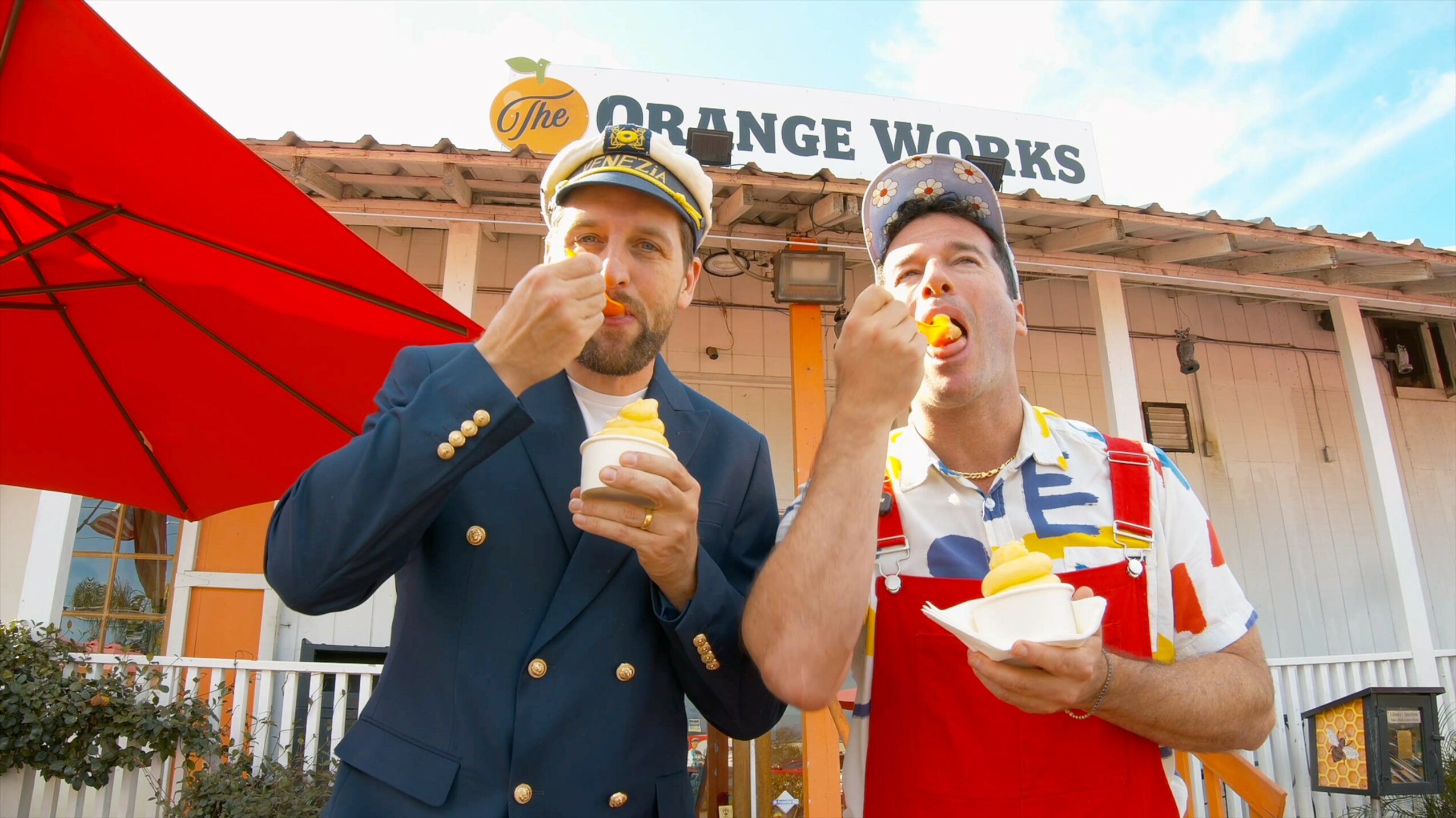 Two men eating ice cream outside a building with a sign reading 'Orange Works', one in a suit and the other in a colorful shirt, with a red umbrella nearby.
