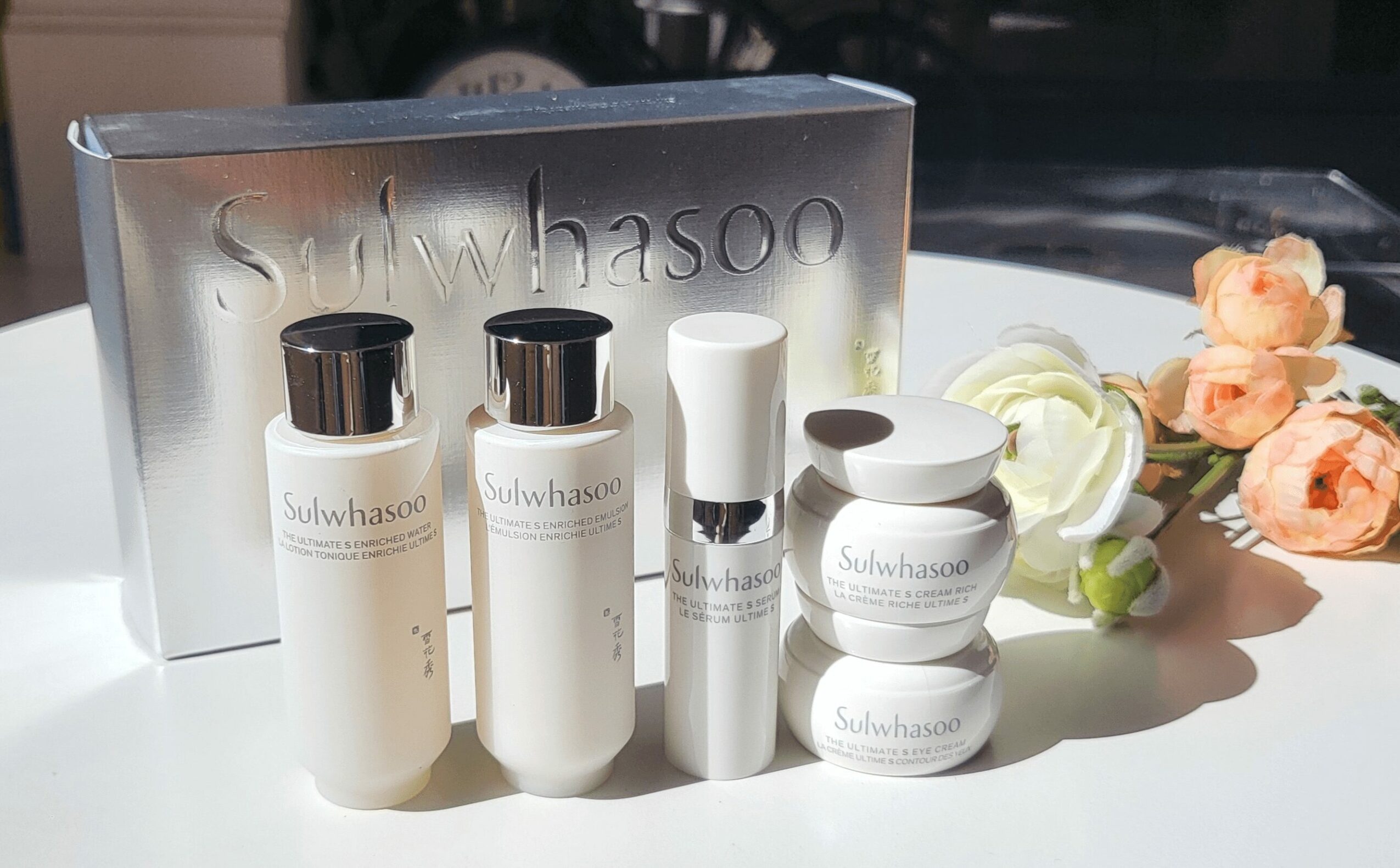 Skincare products in white containers with a silver box, flowers, and a white surface, sunlight casting shadows.