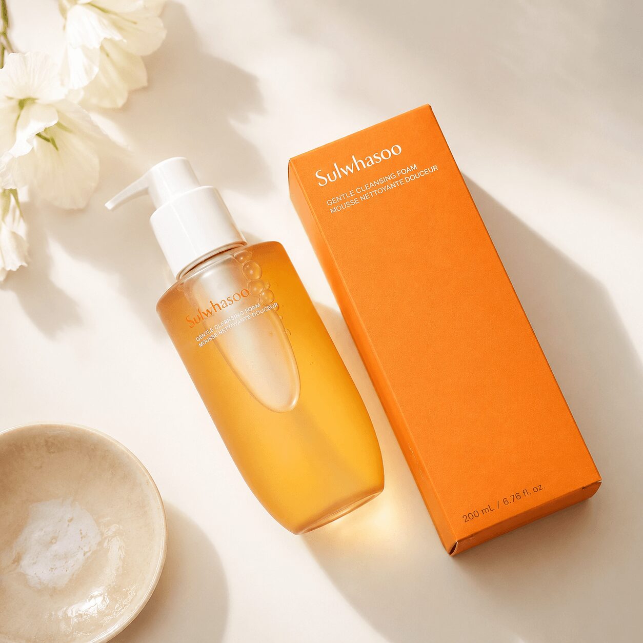 A bottle of Sulwhasoo skincare product and its orange box on a white surface with a bowl and flowers nearby.