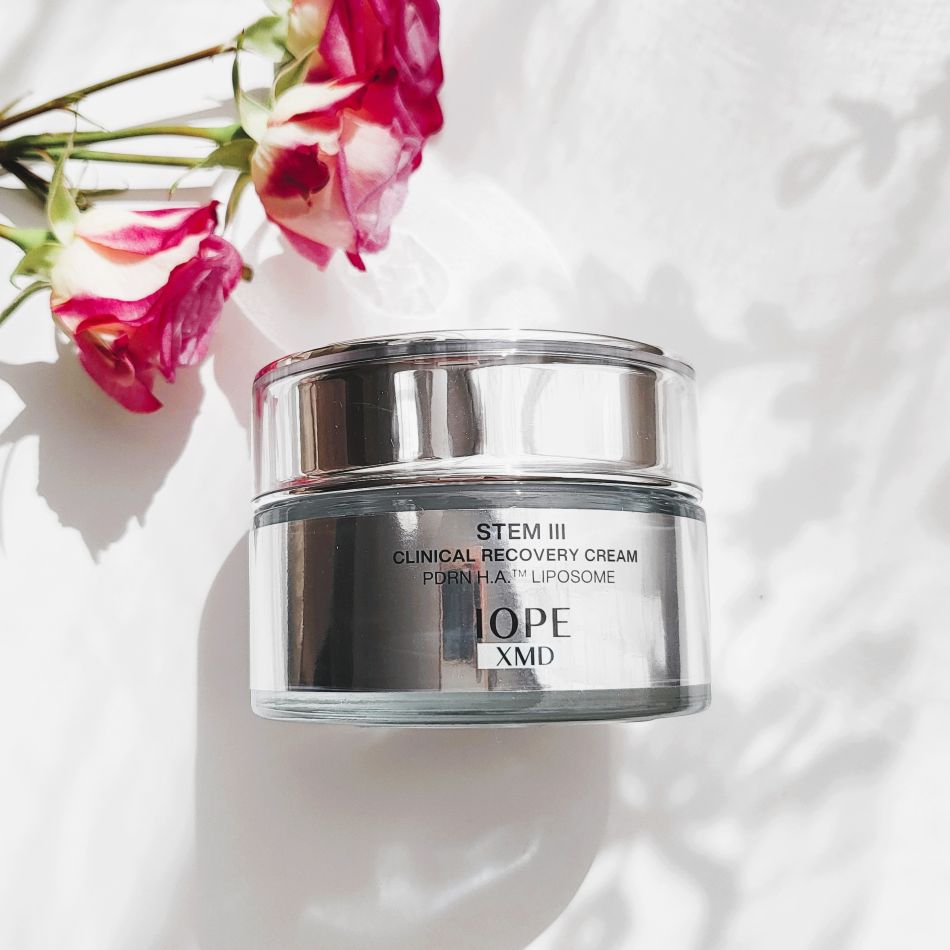 A clear jar of cream with a silver lid, placed on a white surface with pink flowers nearby, shadows cast on the surface.