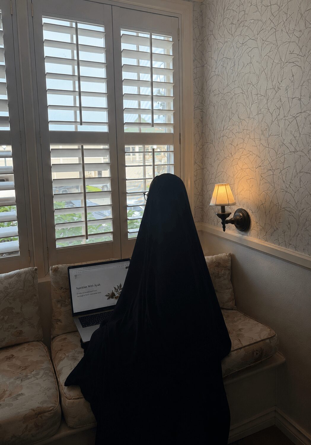 Person wearing a black hijab sitting on a sofa with a laptop, near a window with shutters and a wall-mounted lamp.