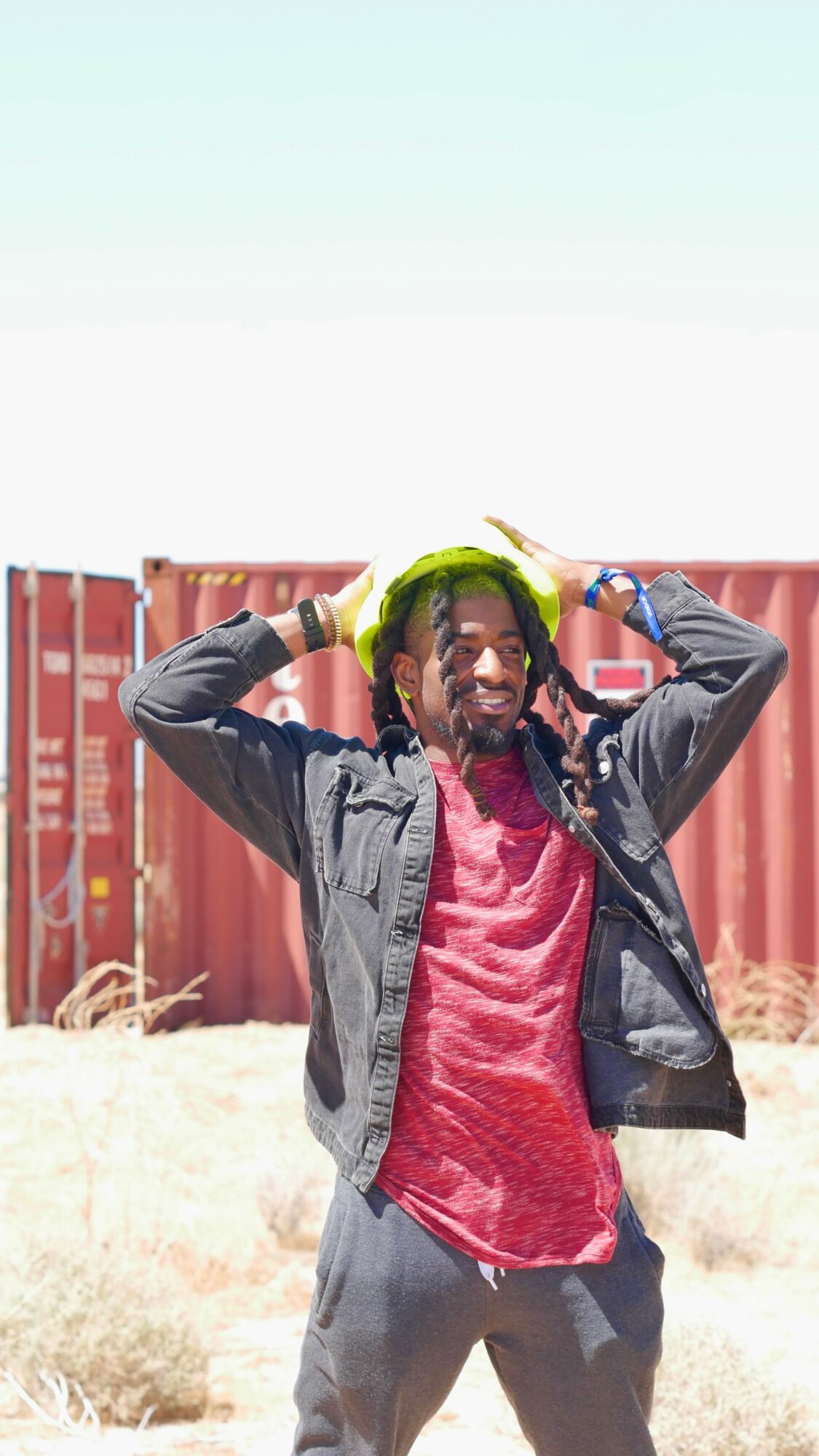 Person with dreadlocks wearing a red shirt and black jacket, smiling outdoors with a red fence in background.