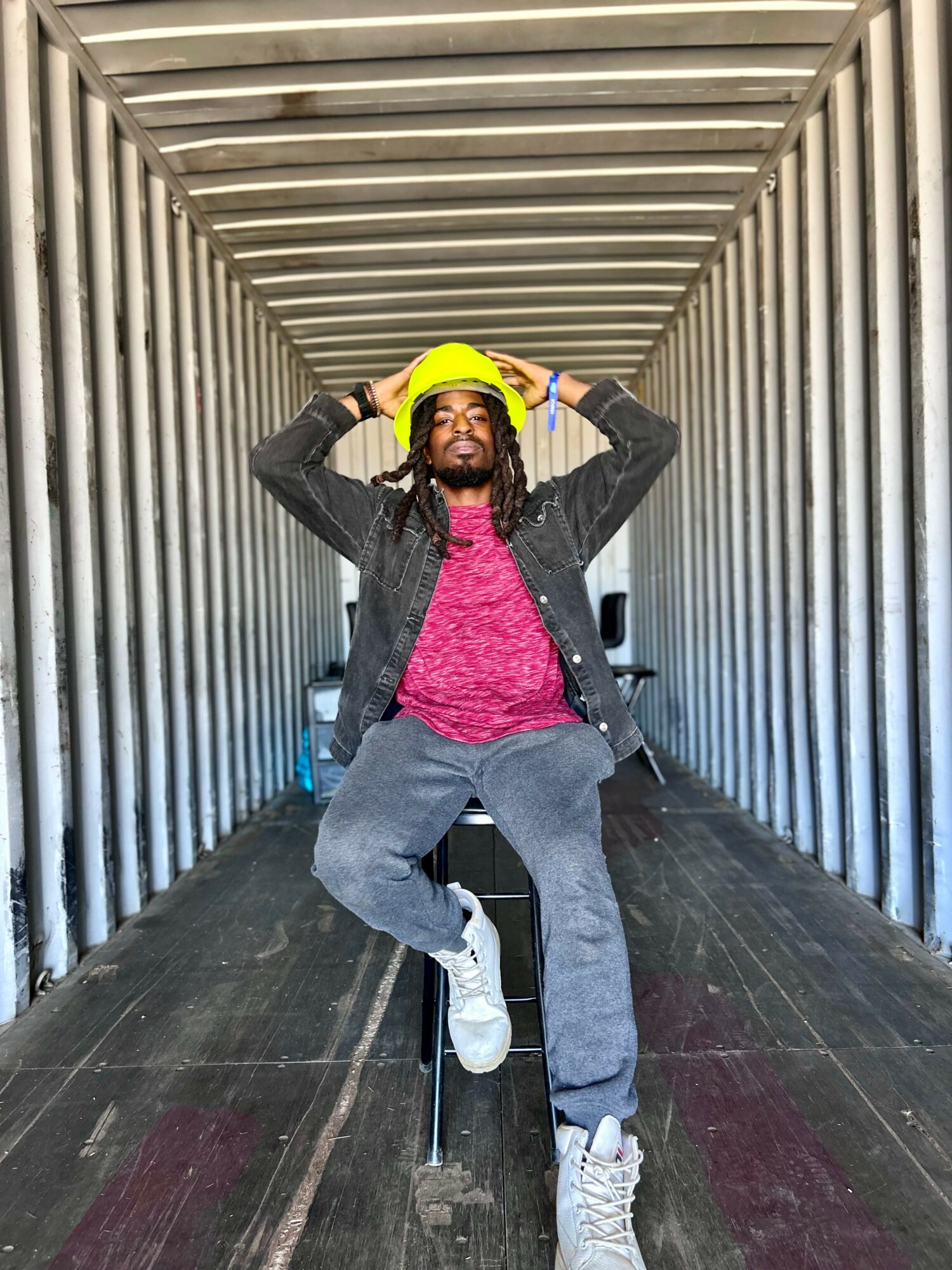 Person sitting in a tunnel with metal walls, wearing a yellow hat, pink shirt, and gray pants, holding the hat with both hands.