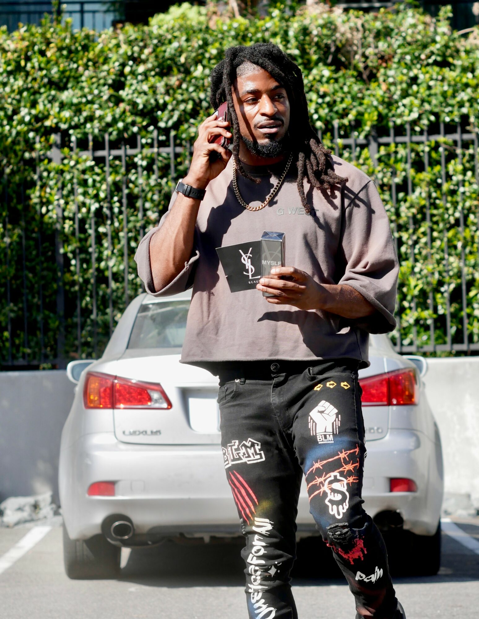 Man with dreadlocks talking on phone, holding a card, standing in parking lot with a silver car and green bushes behind.